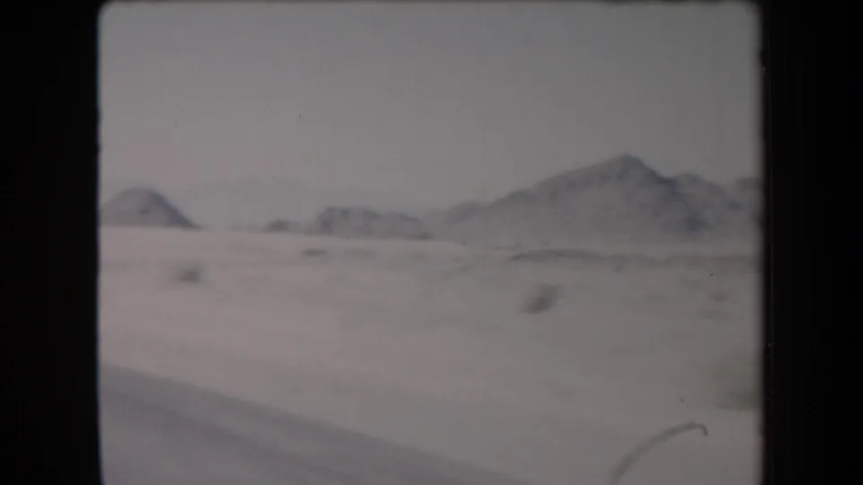 1958 Arizona travel frame with open desert, distant low mountains, hazy sky, and a small trace of the filming vehicle at the lower edge.