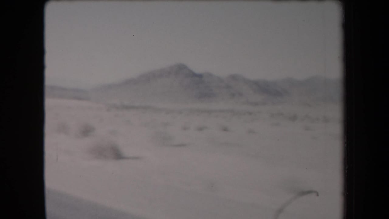 Soft 8mm view of pale desert ground with sparse shrubs and distant low mountains.