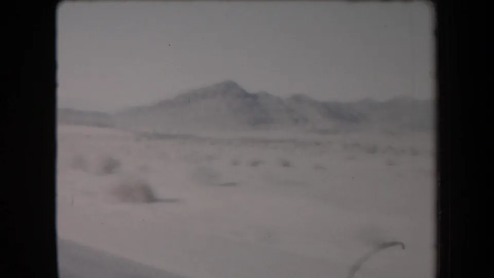 1958 Arizona home-movie frame showing arid desert terrain, scattered brush, and hazy mountain ridges from a moving vehicle.