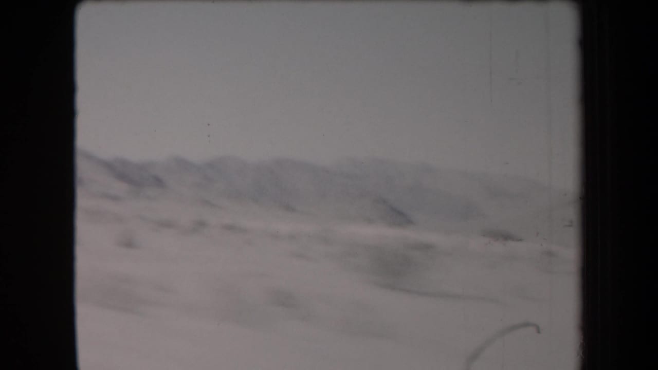1958 Arizona desert road-trip frame with low mountain ridges, open scrubland, and soft motion blur from travel.