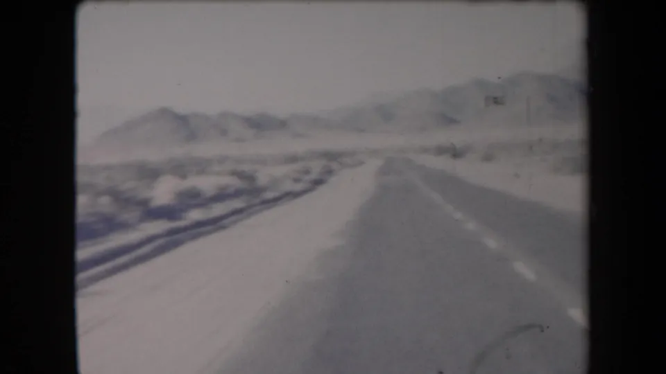 1958 Arizona home-movie frame showing a forward view along a snowy road with low brush and distant mountains.