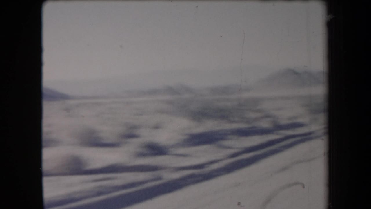 1958 Arizona road-trip frame looking across snow-dusted desert brush toward low mountains from a moving vehicle.