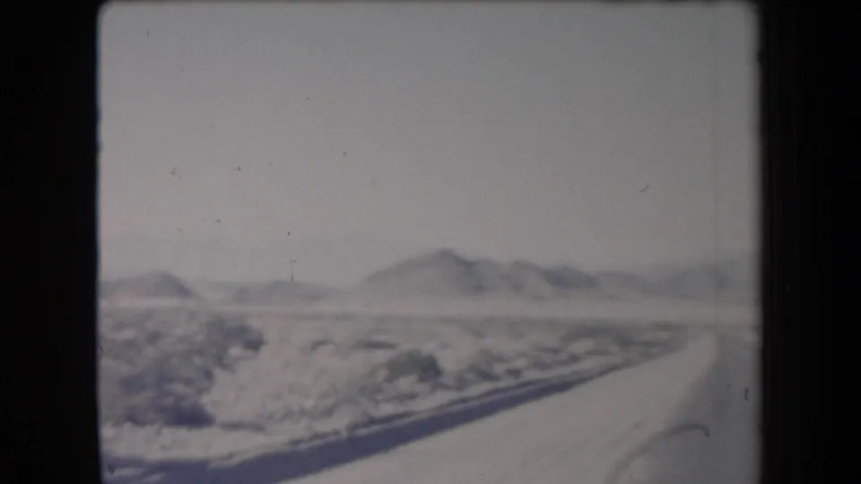 1958 Arizona frame showing a curving road through snow-covered desert brush with low mountains in the distance.