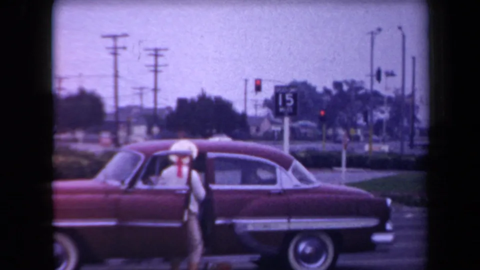 A person in light clothing and a white hat stands next to a red 1950s sedan at a roadside intersection in Arizona.