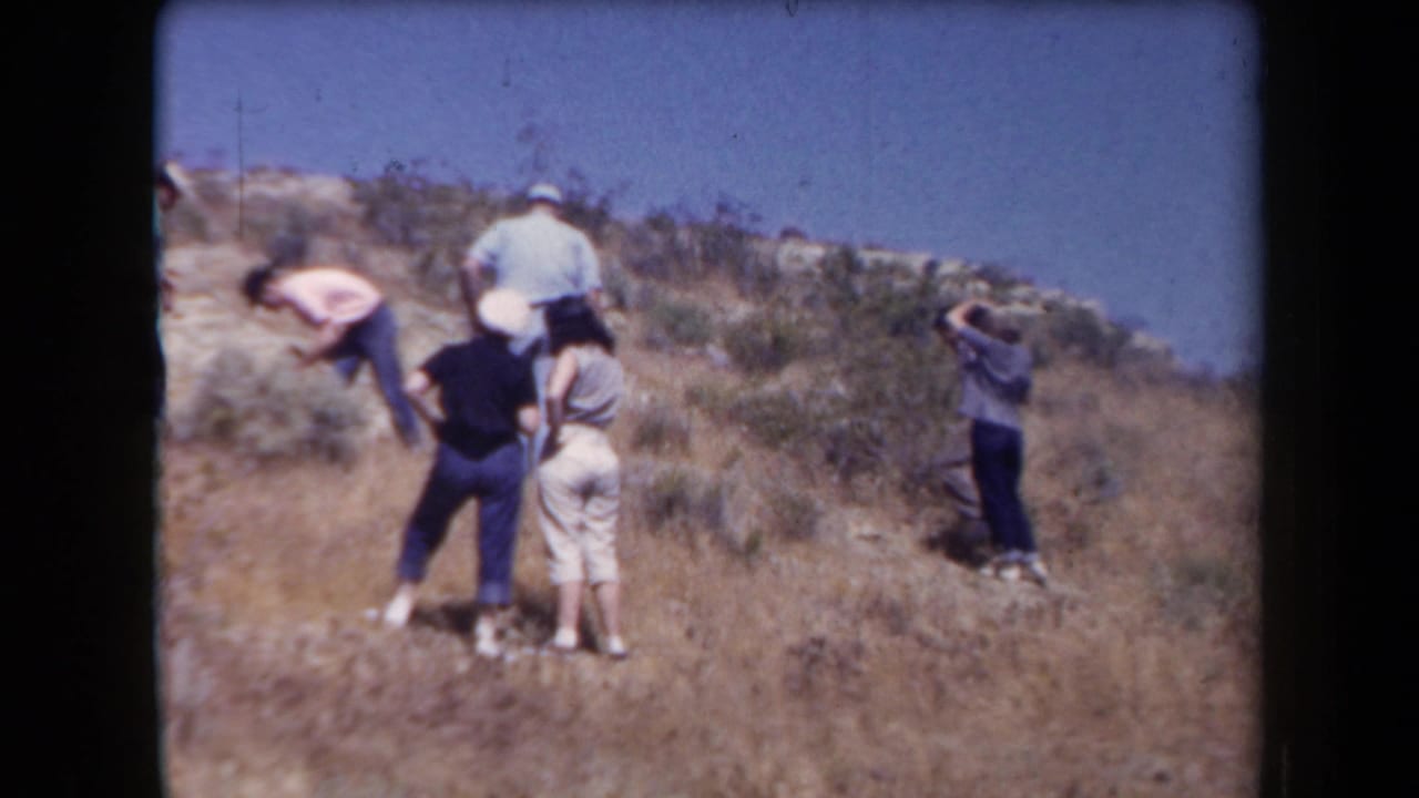 1958 Arizona home-movie frame showing a small group hiking and pausing on a dry, rocky hillside with sparse brush.
