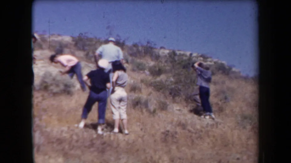 1958 Arizona home-movie frame showing a small group hiking and pausing on a dry, rocky hillside with sparse brush.