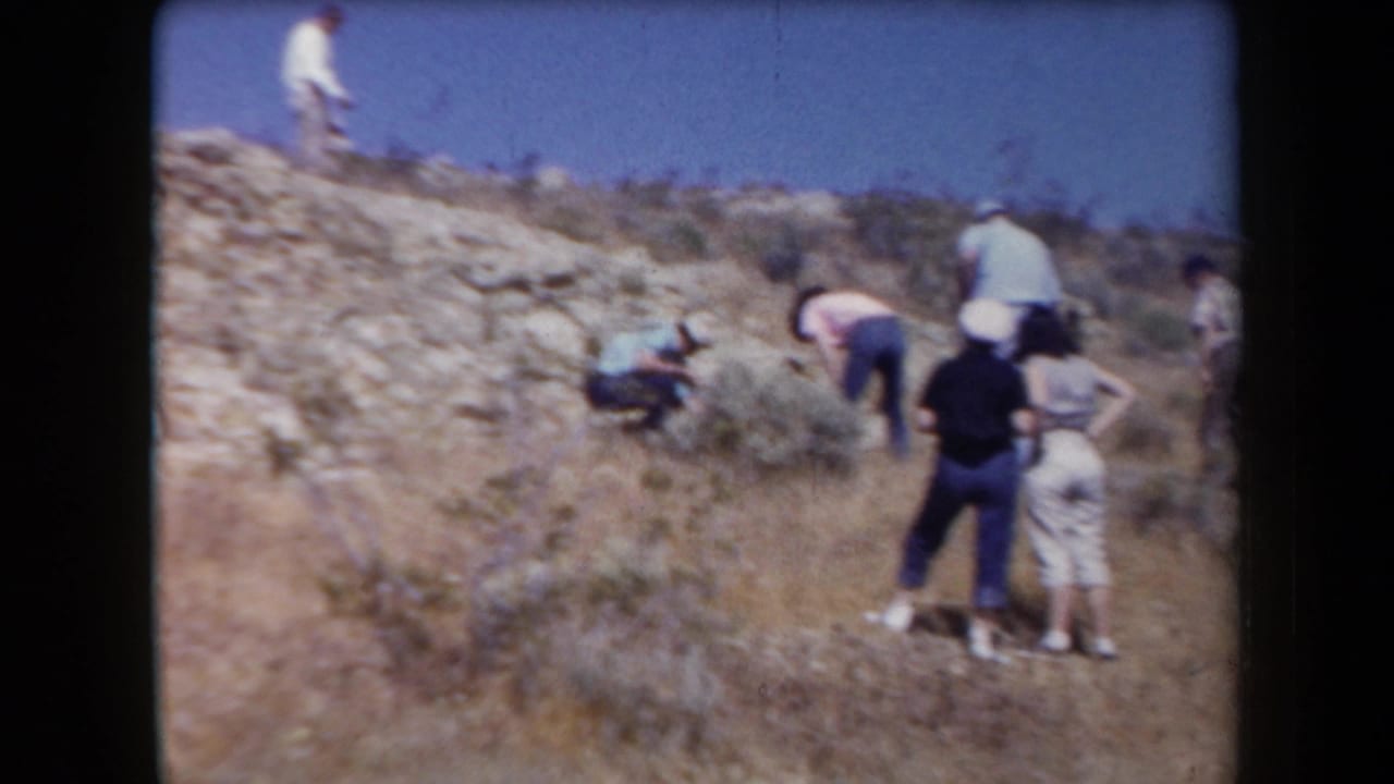 People move across a pale rocky slope with desert brush and open sky behind them.