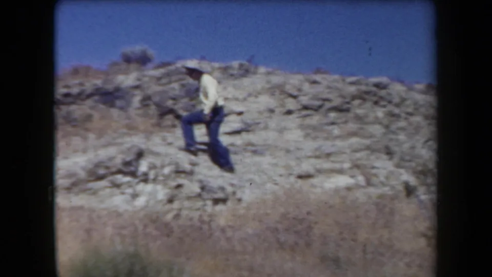 1958 Arizona home-movie frame showing one hiker crossing exposed rock on a dry hillside beneath a blue sky.