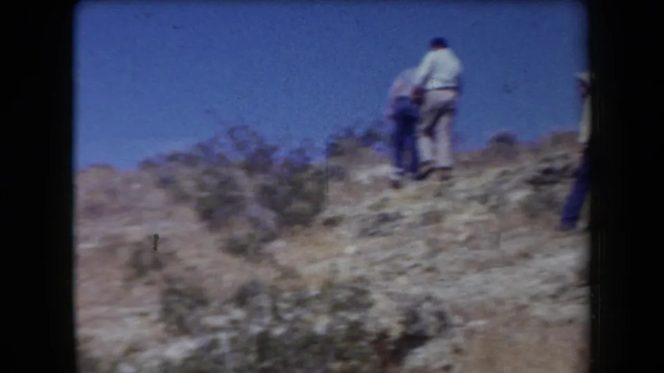 1958 Arizona frame showing hikers near the crest of a dry rocky hillside with brush in the foreground.