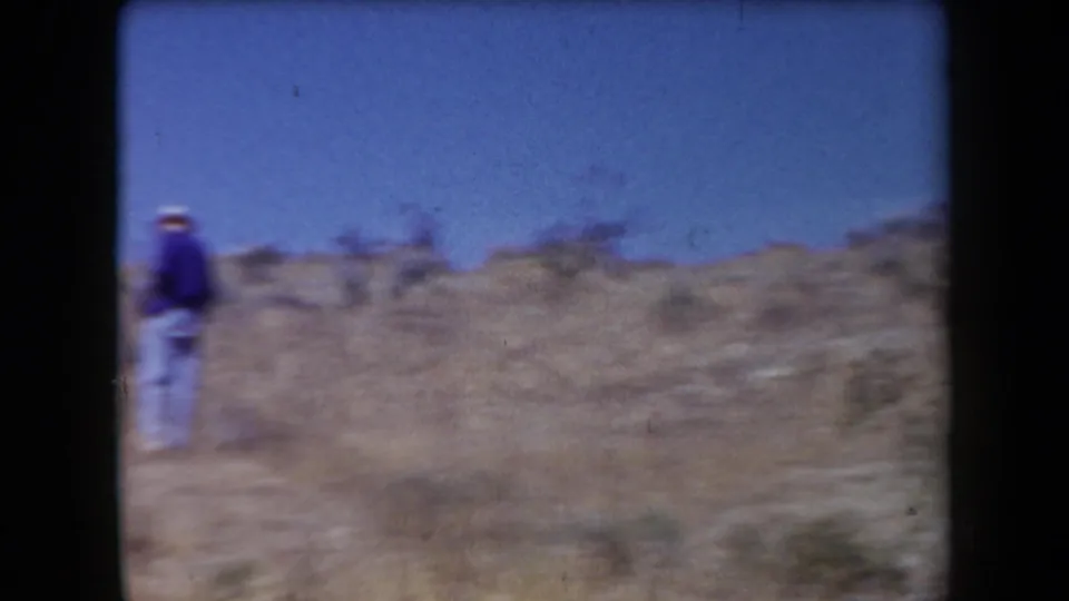 1958 Arizona 8mm frame showing a distant walker on a scrubby desert hillside under clear sky.