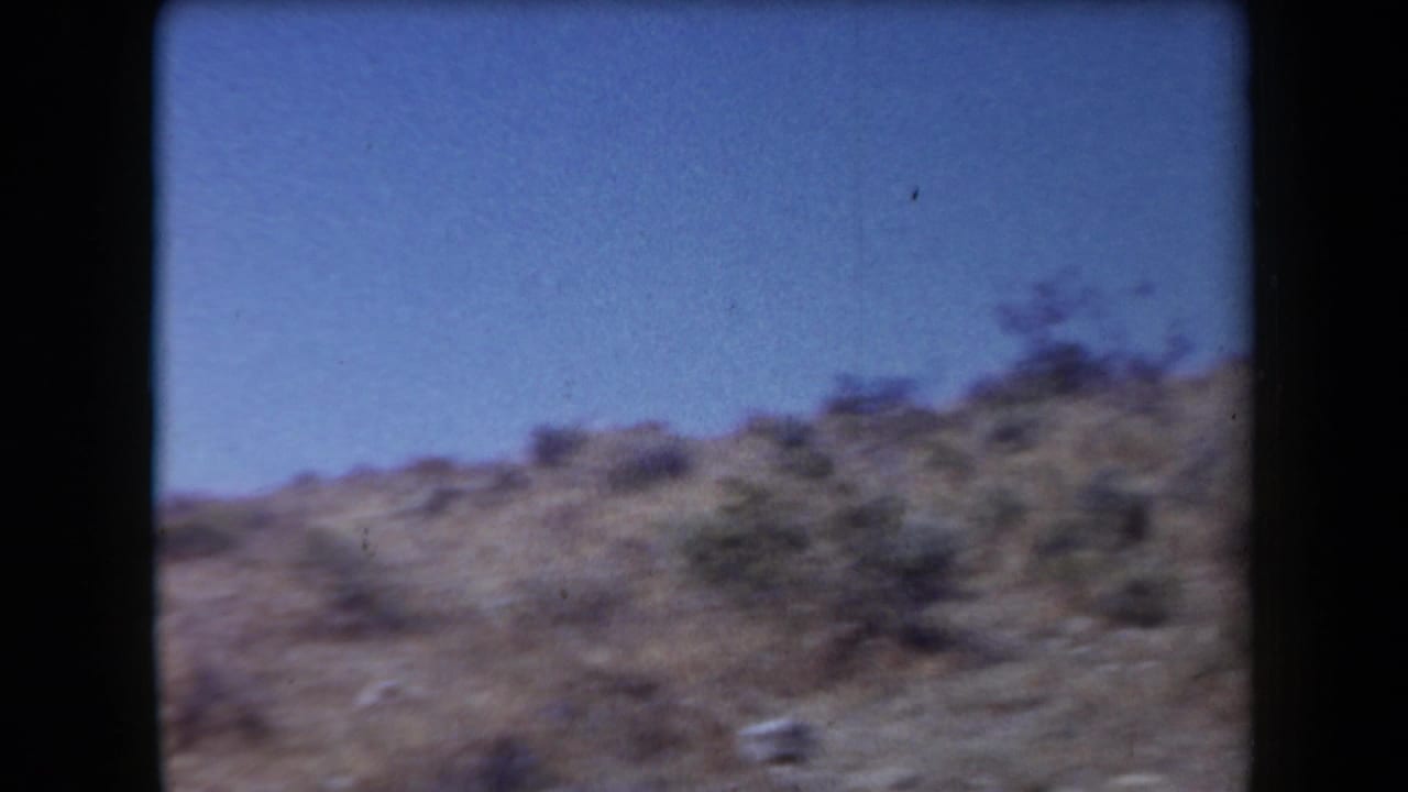 Arizona desert hillside in soft 8mm color, with sparse brush and rocky terrain visible.