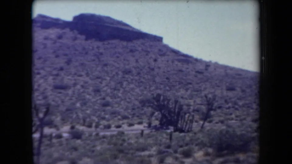 Arizona desert slope with scattered shrubs and a dark ridge, captured in grainy 8mm home-movie footage.