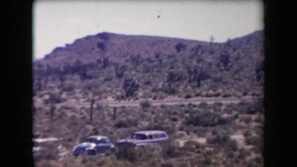 1958 Arizona 8mm frame with two mid-century cars parked near a desert road below rocky hills.
