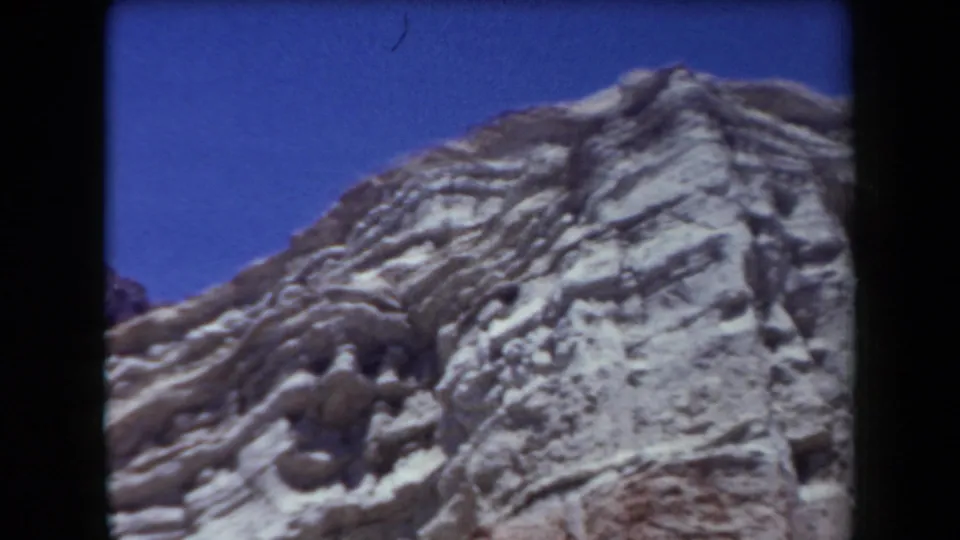 1958 Arizona 8mm frame of a rocky layered ridge and pale slope beneath a deep blue sky.