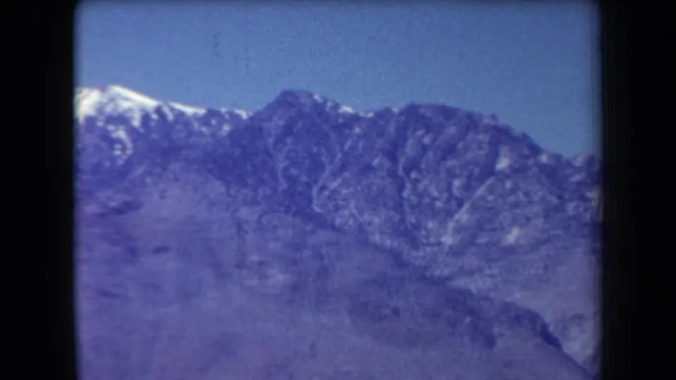 1958 Arizona 8mm frame showing a distant rugged mountain range with one snow-capped peak under blue sky.