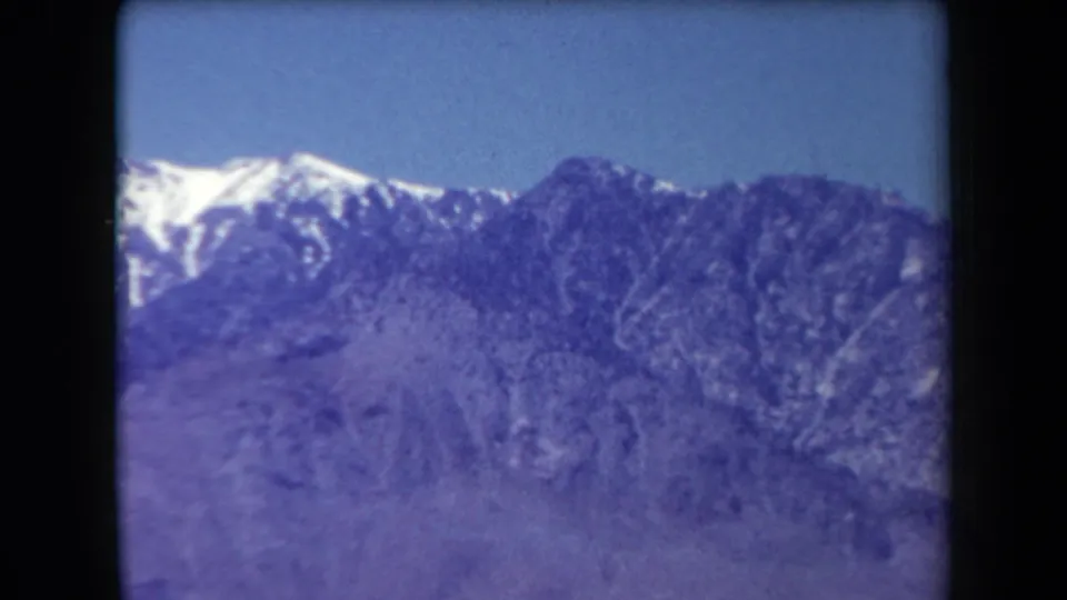 1958 Arizona 8mm frame showing distant snow-covered mountains and rugged slopes beneath a clear blue sky.