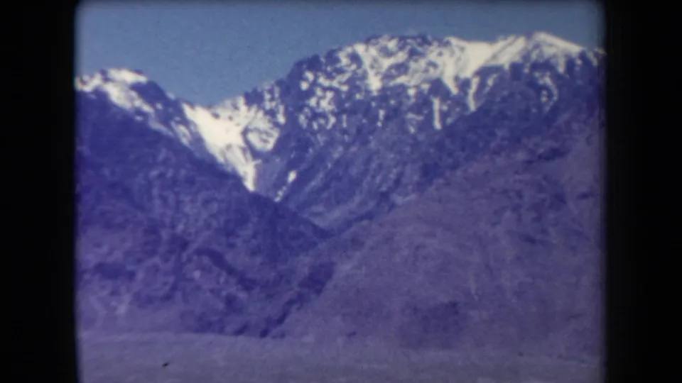 1958 Arizona 8mm frame showing rugged snow-dusted mountain peaks beneath a clear blue sky, with soft focus and purple color shift.