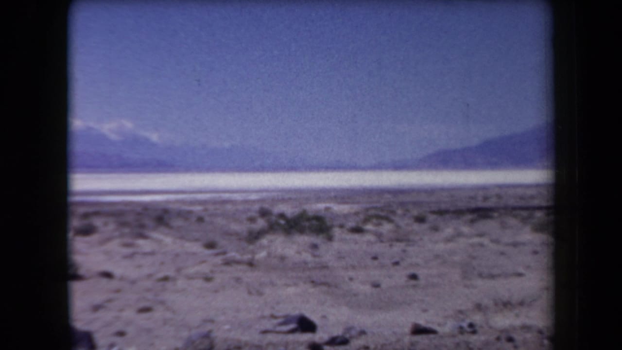 Desert flats with sparse shrubs, distant mountains, and snow-tipped peaks under a blue sky.