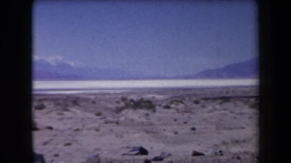 1958 Arizona 8mm frame showing a pale desert foreground, scattered brush, distant ridges, and faint snow-capped mountains.