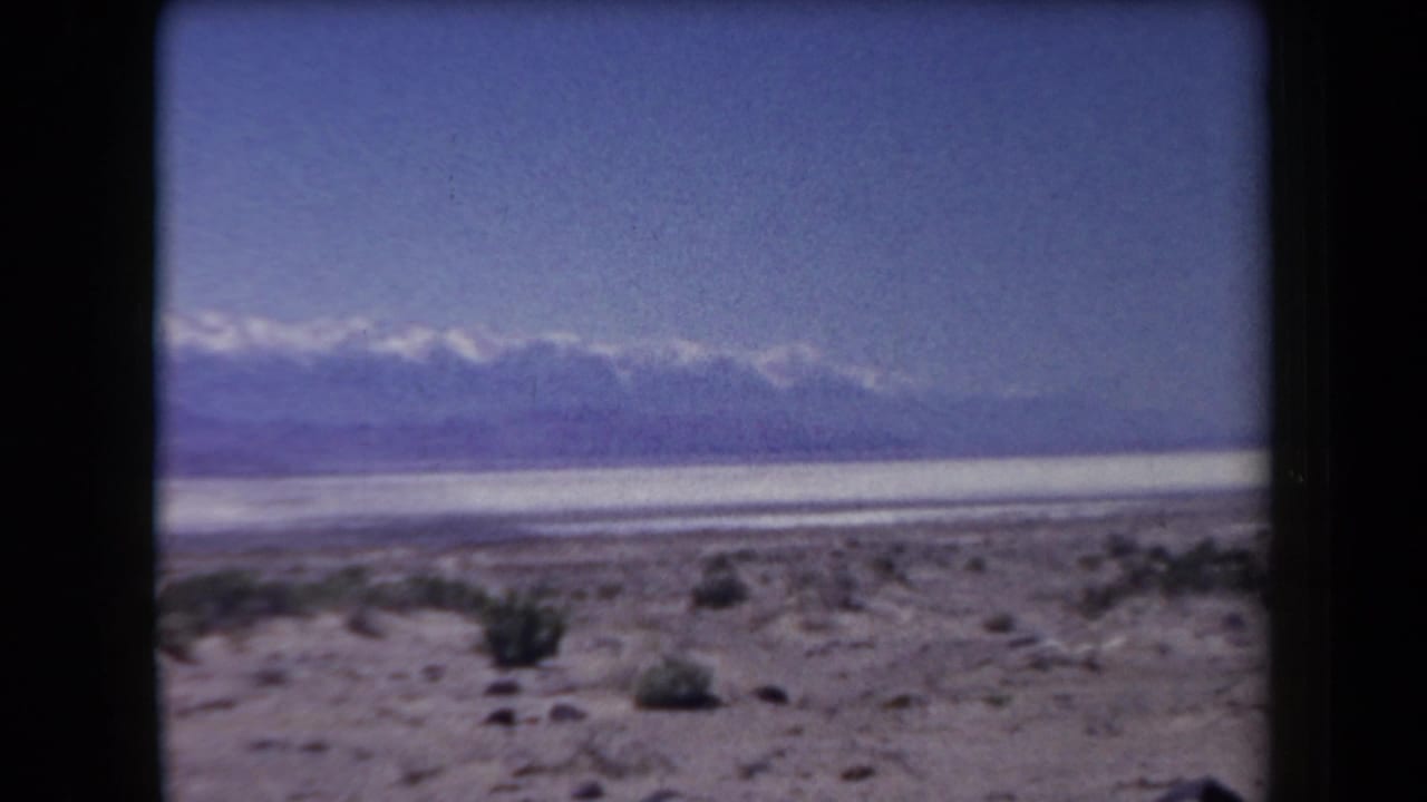 Open desert floor with brush, a pale flat basin, and snow-capped mountains in the distance.
