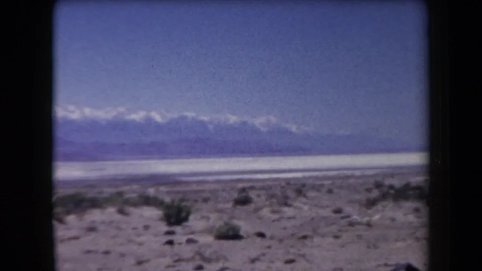 1958 Arizona home-movie frame looking across sparse desert vegetation toward a light basin and distant snowy mountain range.