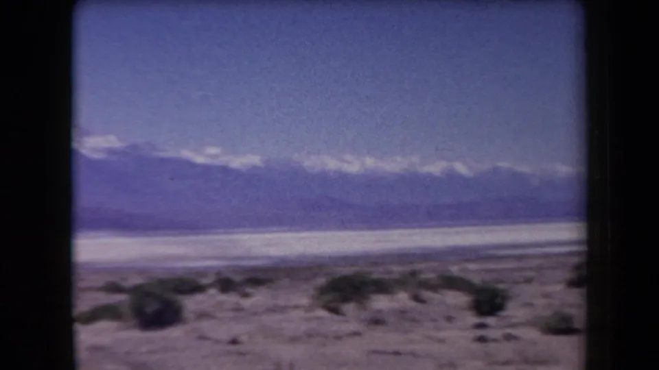 1958 Arizona 8mm view with dark foreground rocks, scattered shrubs, a pale salt-flat-like basin, and hazy mountain ridges.