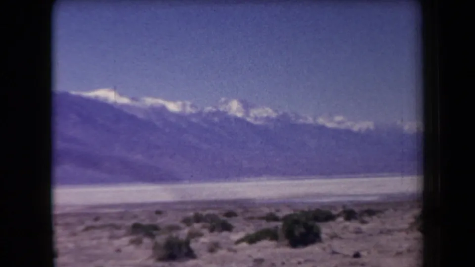 1958 Arizona 8mm frame with a closer dark mountain ridge, snowy peaks beyond, and sparse desert vegetation in front.