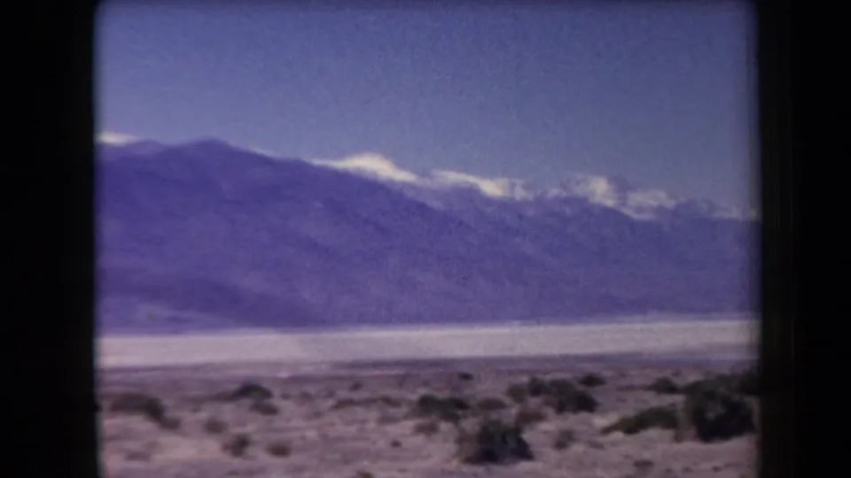 1958 Arizona 8mm frame showing sparse desert scrub, pale flats, and distant mountains with small snow patches beneath a blue sky.