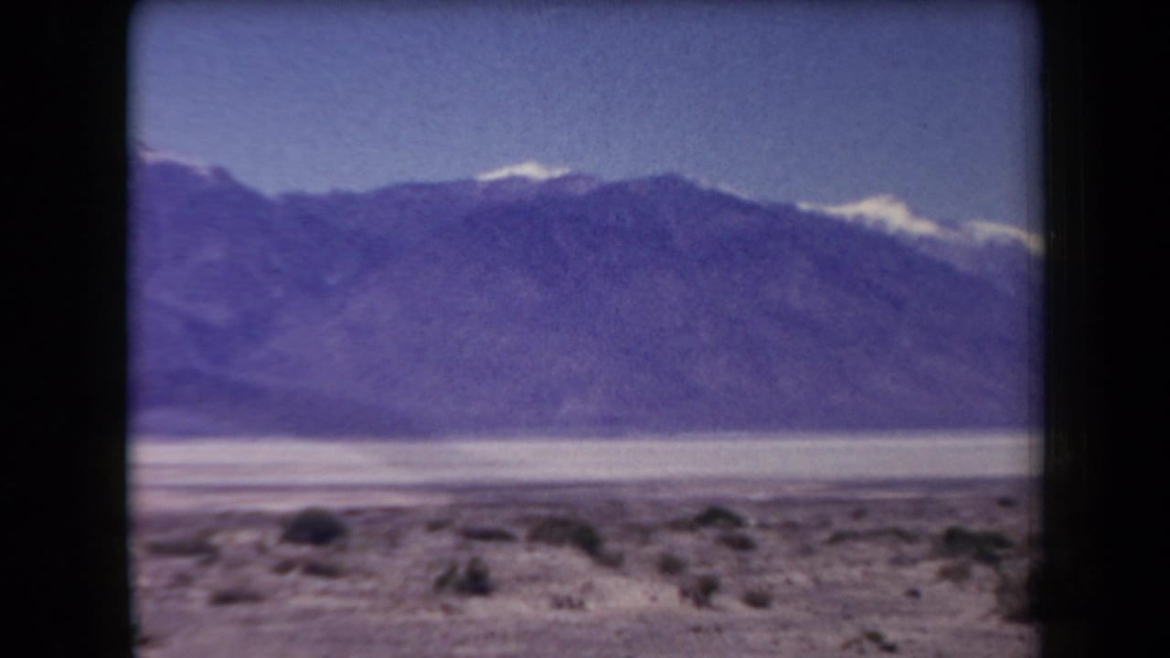 1958 Arizona home-movie frame with a broad mountain ridge, bright desert basin, sparse foreground shrubs, and faded 8mm color.