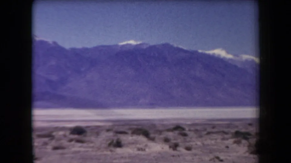 1958 Arizona home-movie frame with a broad mountain ridge, bright desert basin, sparse foreground shrubs, and faded 8mm color.