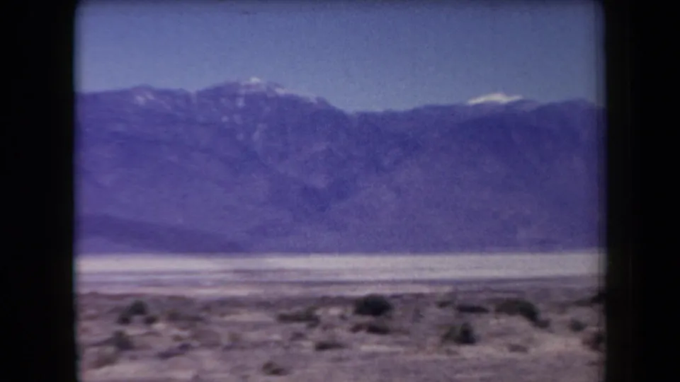 1958 Arizona 8mm frame showing a scrub-covered foreground, pale desert floor, and dark mountain range with faint snow highlights.