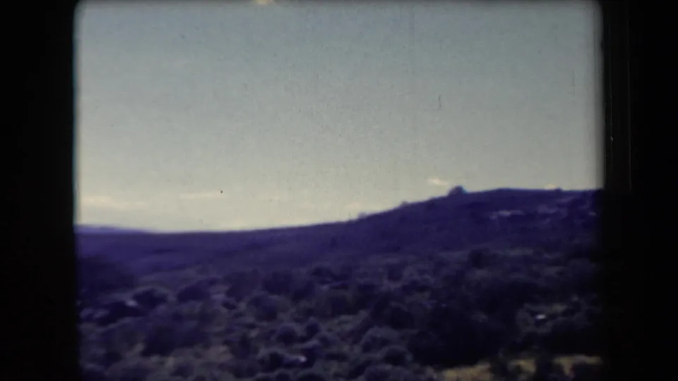 1958 Arizona 8mm frame showing a darker scrubby hillside or ridge line under a pale sky, with heavy shadow and film-edge darkness.
