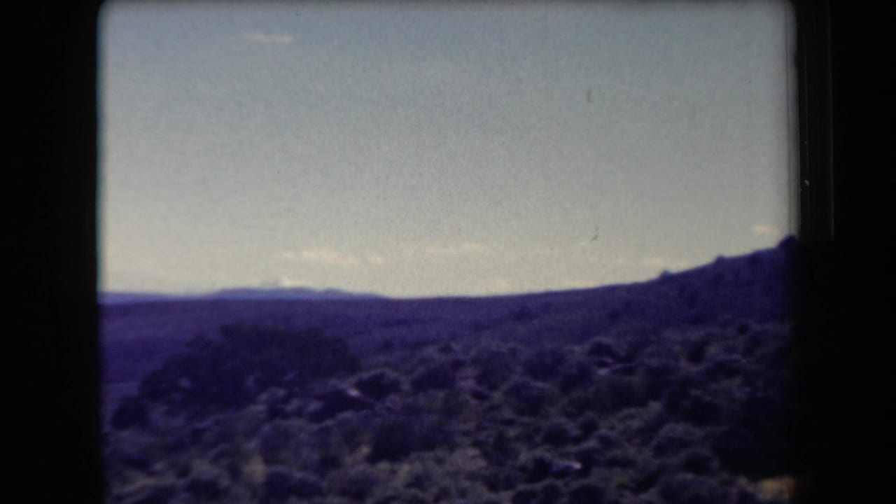1958 Arizona 8mm frame showing scrub-covered desert hills and distant mountain ridges beneath a pale sky.