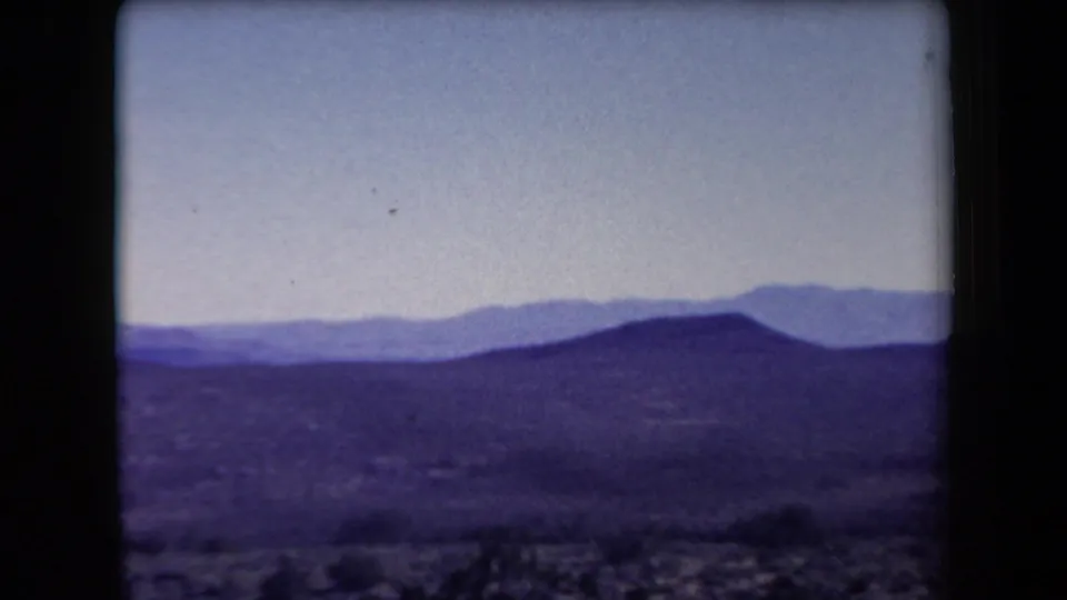 1958 Arizona 8mm frame with a low desert ridge, distant mountains, pale sky, and soft purple-tinted foreground terrain.