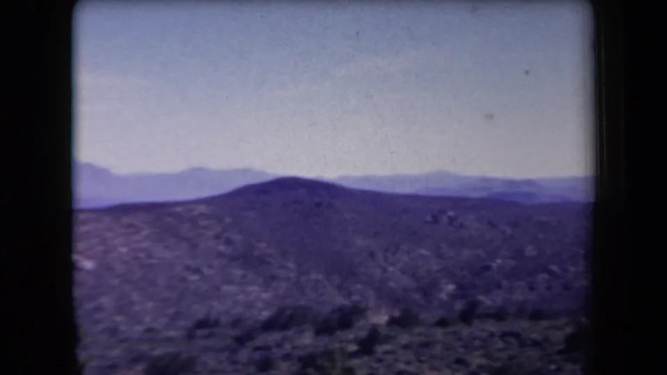 1958 Arizona home-movie frame showing a rounded desert hill, sparse scrub, distant mountains, and a pale blue sky.