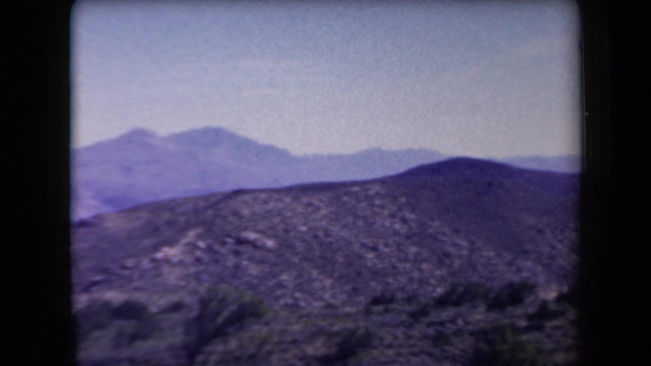 Rocky desert hills in front of distant mountain ridges under a pale sky.