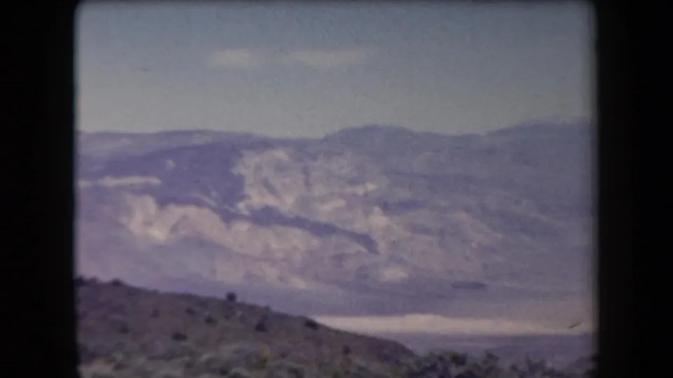 1958 Arizona 8mm frame looking over a low hillside and small tree toward a hazy valley and distant mountain ridges.
