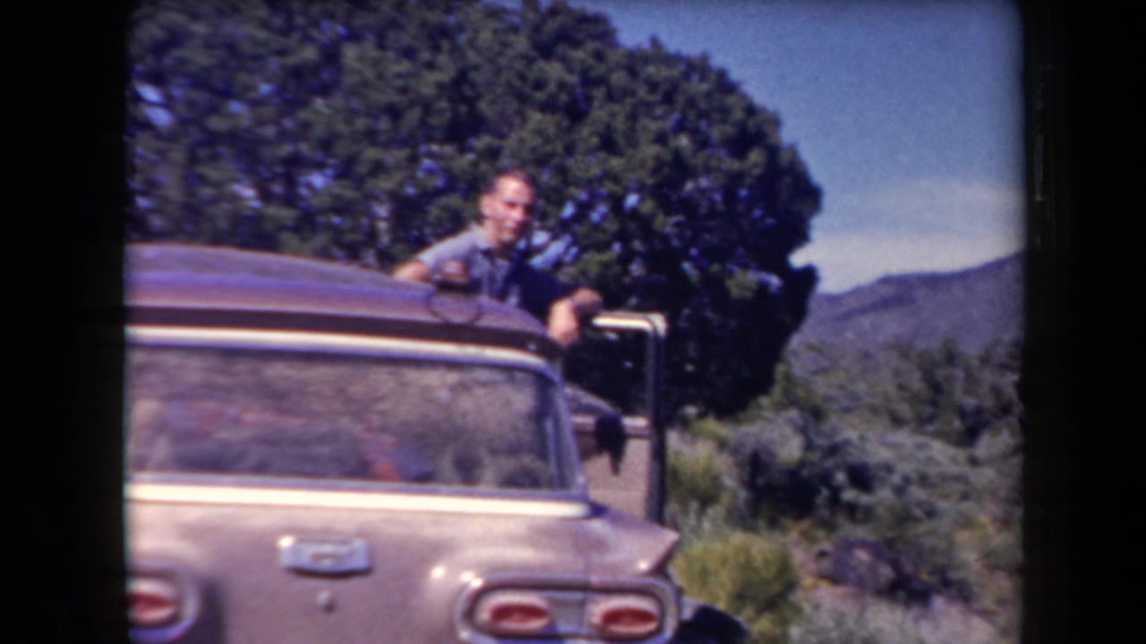 Arizona 1958 home-movie frame with an adult man leaning from a station wagon beside desert vegetation and distant mountains.