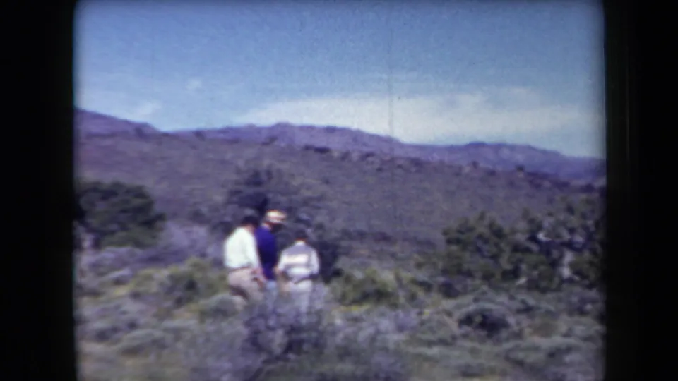 Arizona 1958 home-movie frame showing two adult men moving through scrubby desert terrain beneath distant hills.