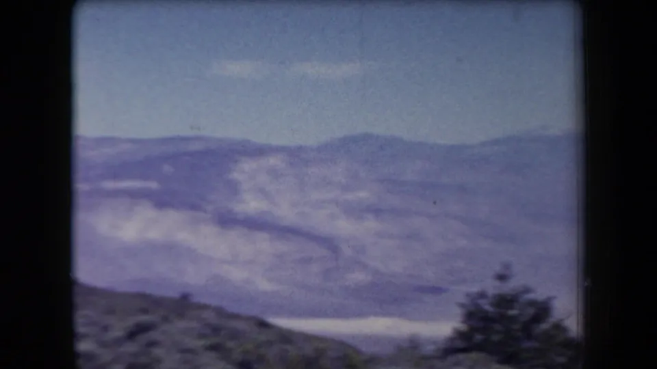 1958 Arizona home-movie frame of layered blue mountain ridges, a pale low area below, and an empty foreground slope.