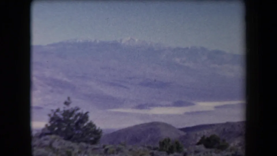 1958 Arizona 8mm frame with dark shrubs or trees, low hills, hazy distant mountains, and a pale water-like surface.