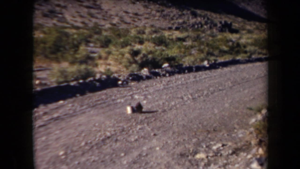 1958 Arizona 8mm frame showing a gravel road, rocky slope, sparse shrubs, and two indistinct objects on the road.