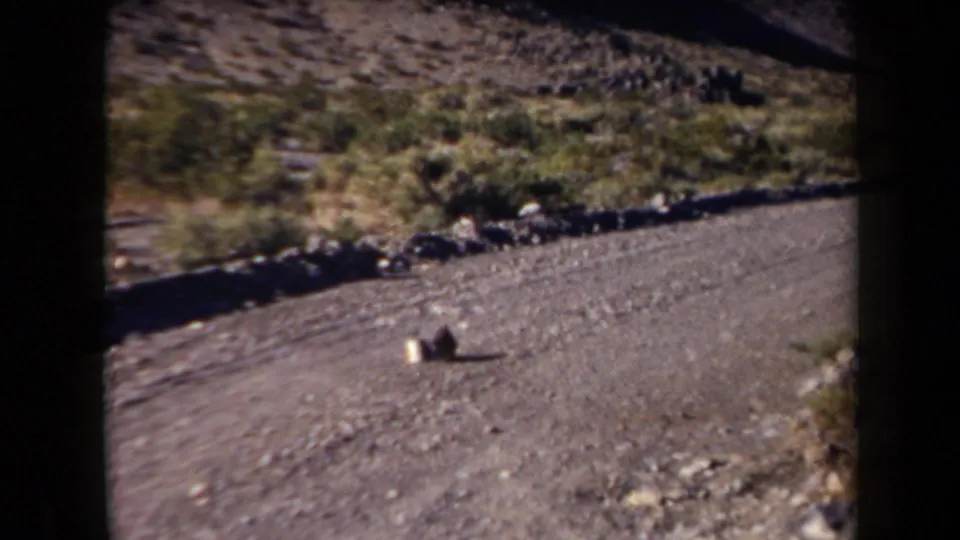 1958 Arizona 8mm frame showing a gravel road, rocky slope, sparse shrubs, and two indistinct objects on the road.