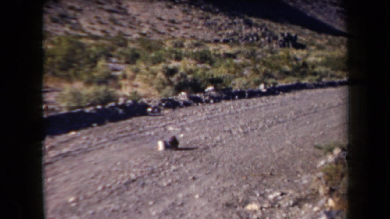 1958 Arizona roadside frame with a gravel surface, scrubby hillside, rocks, and two indistinct objects in view.