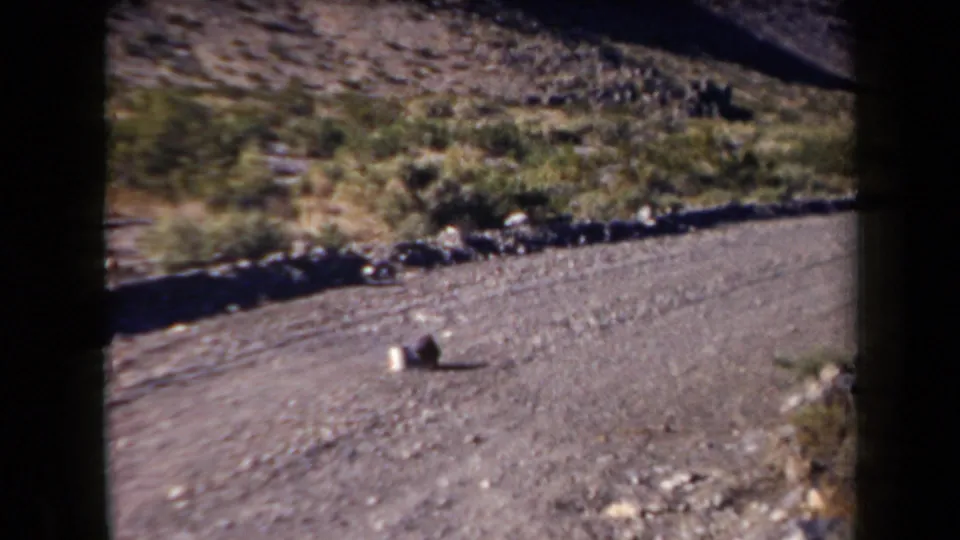1958 Arizona roadside frame with a gravel surface, scrubby hillside, rocks, and two indistinct objects in view.