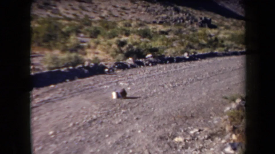 Arizona home-movie frame from 1958 showing an angled gravel road, dry hillside, shrubs, and two small road objects.