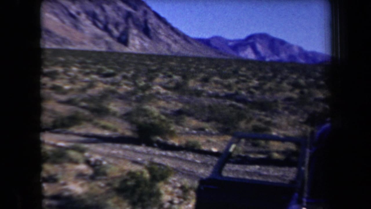 Rocky desert mountains and brush seen beside a dark car door or frame edge.