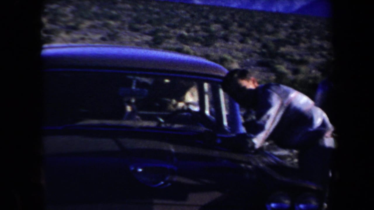 An adult man leans over the hood area of a dark mid-century car in the Arizona desert landscape.