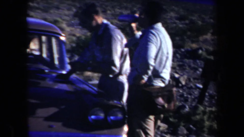 Three adult men in casual shirts gather by the front of a dark car during a 1958 Arizona roadside stop.