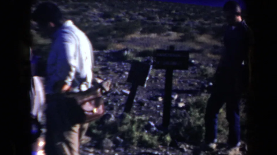 Adult men stand near brush and a signpost-like object beside the stopped car in Arizona home-movie footage.